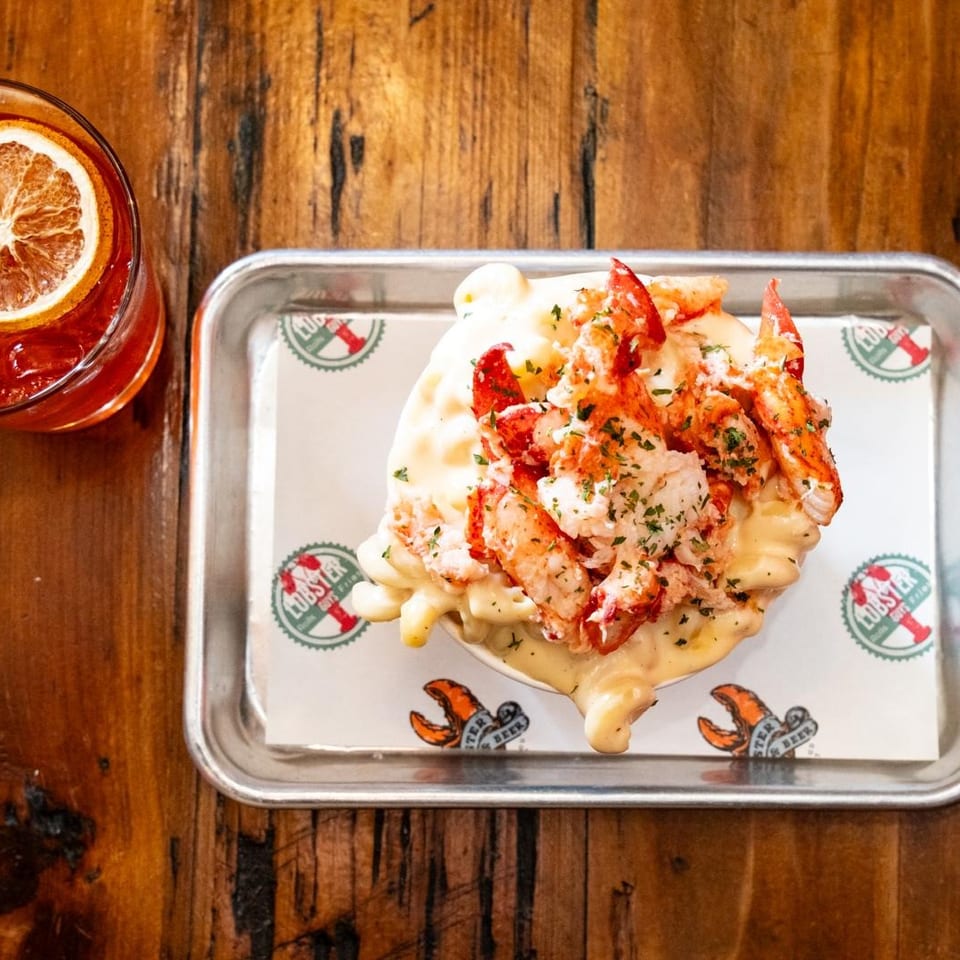 Lobster mac and cheese on a tray with a red cocktail garnished with an orange slice.
