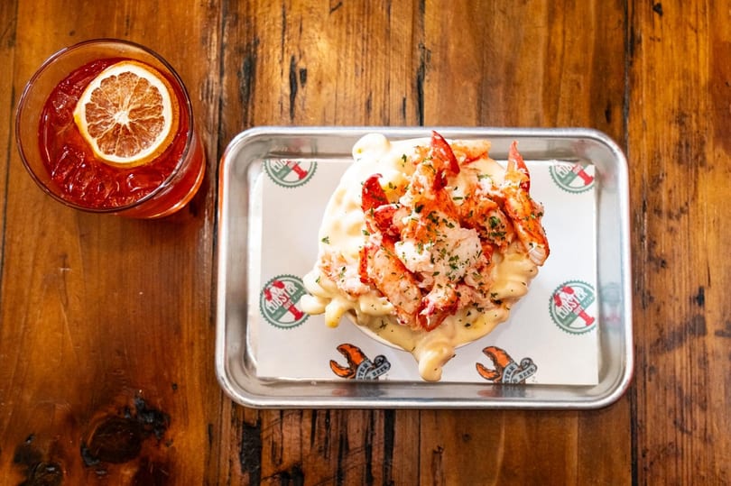 Lobster mac and cheese on a tray with a red cocktail garnished with an orange slice.