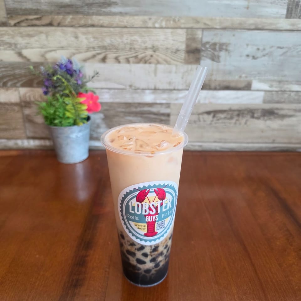 Iced milk tea with tapioca pearls and a straw, branded Lobster Guys, sits on a wooden table beside a small flower pot.