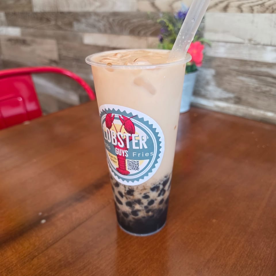 A creamy bubble tea with tapioca pearls and a Lobster Guys Fries logo sits on a wooden restaurant table.