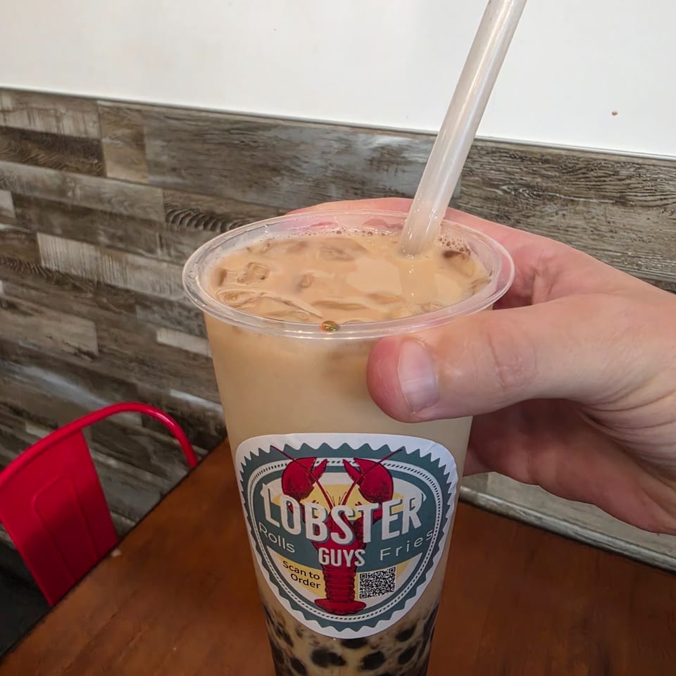 Hand holding a Lobster Guys bubble tea with tapioca pearls, creamy iced drink, straw, and branded cup on a wooden table.