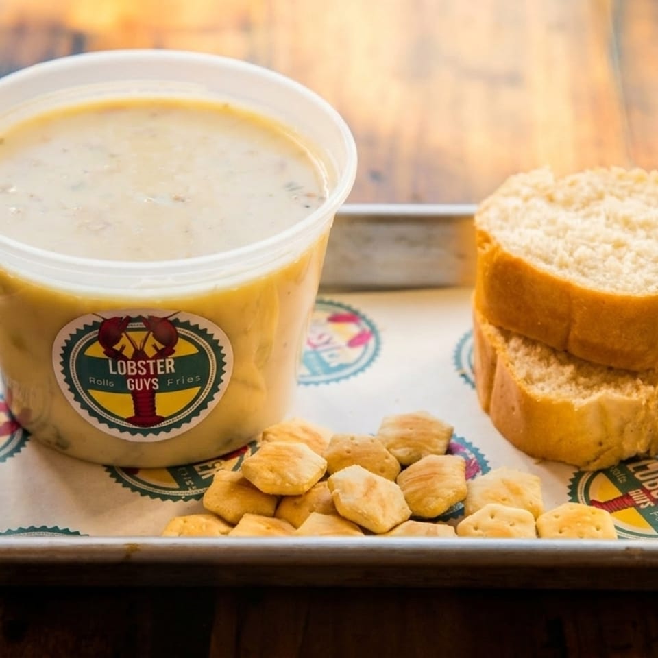 Lobster Guys clam chowder in a plastic container with bread slices and crackers on a tray.