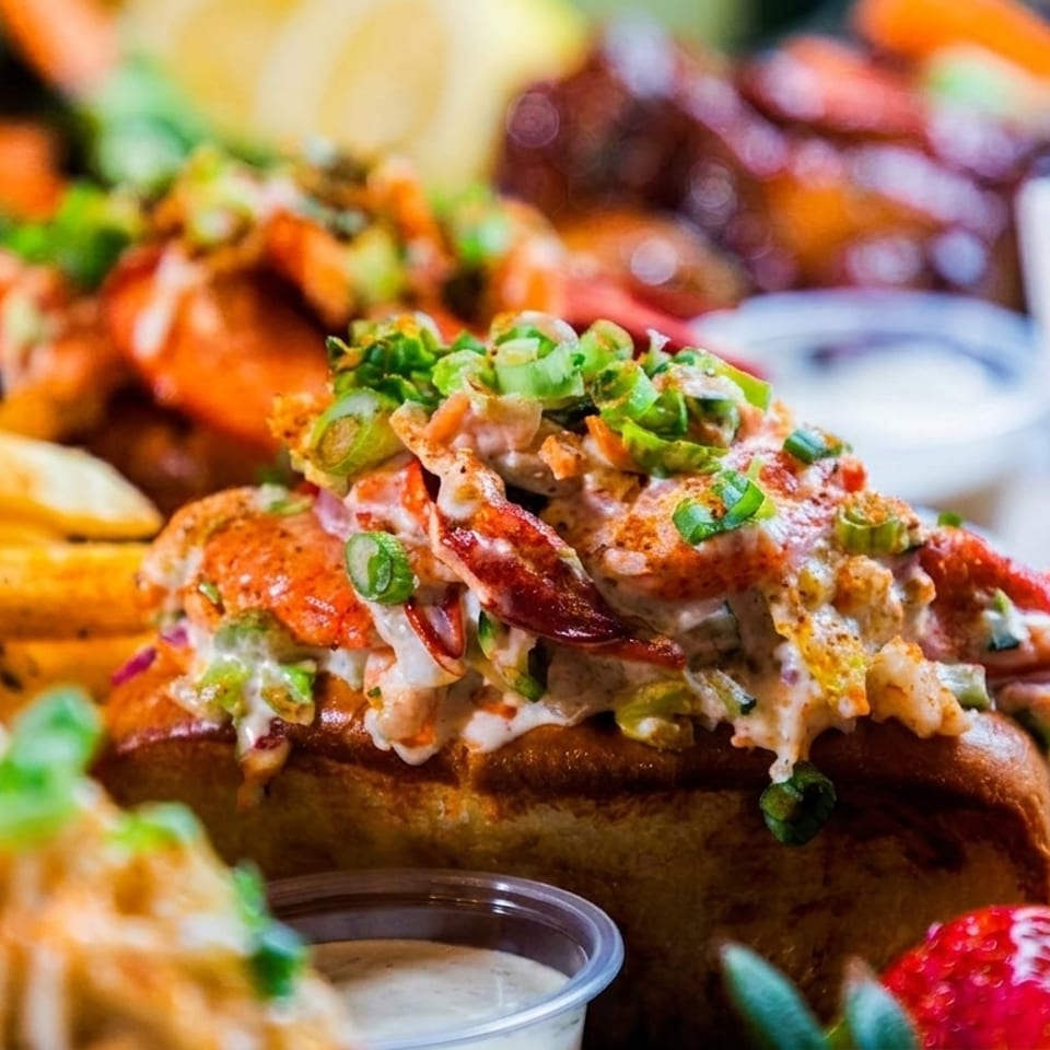 Close-up of a lobster roll topped with green onions, with fries and a drink in the background.